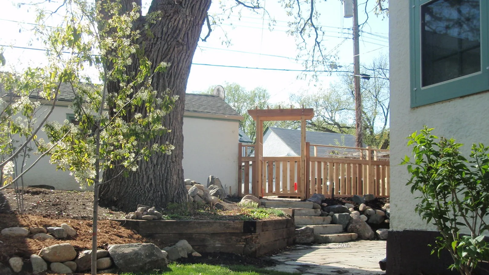 A backyard has separate lawn sections, a tree, stone borders, steps, and a gate.